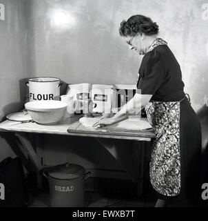 Historische, 1950er Jahre, auf einem kleinen Marmor tisch in einer Ecke des Raumes, eine reife Lady das Tragen einer Schürze Gebäck, mit einem Holz- Rolling Pin auf ein Schneidebrett zu glätten und die Form der dougn vor dem Backen. Stockfoto