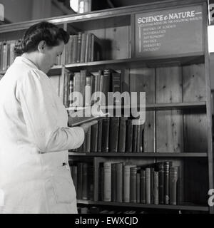 1950er Jahre, historisch, eine Dame in einem weißen Mantel, Stift in der Seitentasche, in einer öffentlichen Bibliothek, stehend und mit Blick auf ein Buch in der Science & Nature Sektion. Stockfoto