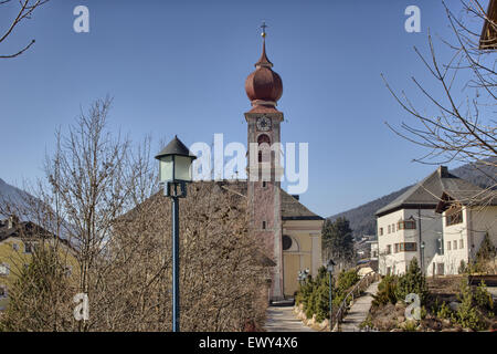 Der Luis Trenker Promenade führt bis St. Ulrich, italienisches Dorf in Dolomiten Alpen: typische Häuser, Gebäude und die roten Glockenturm mit Uhr Pfarrkirche St. Ulrich mit schneebedeckten Bergen und grünen Koniferen im Hintergrund Stockfoto