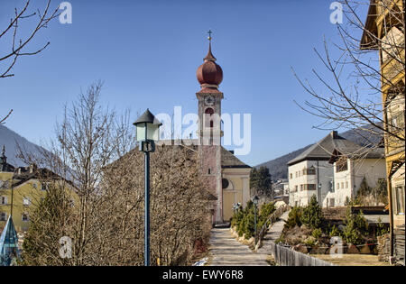 Der Luis Trenker Promenade führt bis St. Ulrich, italienisches Dorf in Dolomiten Alpen: typische Häuser, Gebäude und die roten Glockenturm mit Uhr Pfarrkirche St. Ulrich mit schneebedeckten Bergen und grünen Koniferen im Hintergrund Stockfoto