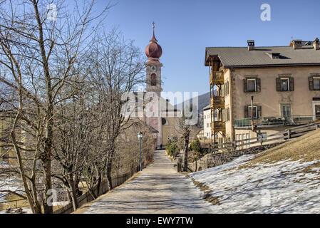 Der Luis Trenker Promenade führt bis St. Ulrich, italienisches Dorf in Dolomiten Alpen: typische Häuser, Gebäude und die roten Glockenturm mit Uhr Pfarrkirche St. Ulrich mit schneebedeckten Bergen und grünen Koniferen im Hintergrund Stockfoto