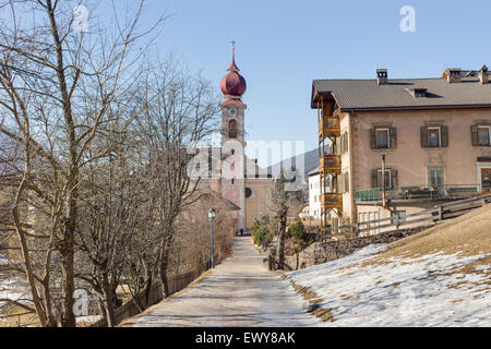 Der Luis Trenker Promenade führt bis St. Ulrich, italienisches Dorf in Dolomiten Alpen: typische Häuser, Gebäude und die roten Glockenturm mit Uhr Pfarrkirche St. Ulrich mit schneebedeckten Bergen und grünen Koniferen im Hintergrund Stockfoto