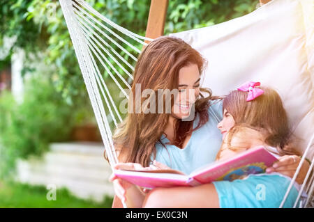 Fröhliche Mutter mit Tochter Spaß im Freien, in der Hängematte entspannen und lesen lustige Geschichte, glückliche Familie Zeit zu verbringen Stockfoto