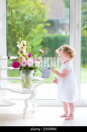 Entzückende lockige Babymädchen in einem weißen Kleid Blumen in einem schönen Wohnzimmer mit großem Fenster und Tür zum Garten gießen Stockfoto