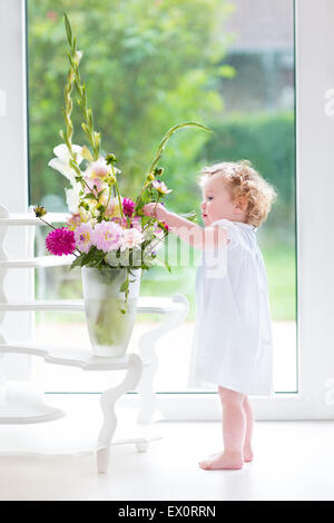 Entzückende lockige Babymädchen in einem weißen Kleid Blumen in einem schönen Wohnzimmer mit großem Fenster und Tür zum Garten gießen Stockfoto