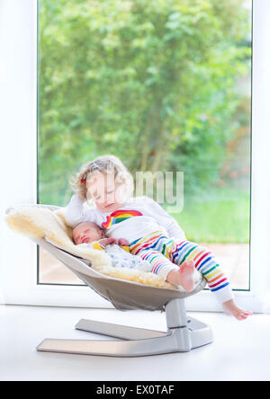 Neugeborenes Baby junge und seine Kleinkind Schwester zum Entspannen in einer Schaukel neben einem großen Fenster und Tür zum Garten Stockfoto