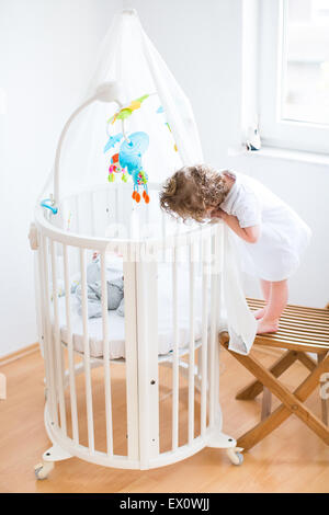 Lustige lockiges Kleinkind Mädchen Blick auf ihr neugeborenes Baby Bruder schlafen in einer weißen runden Krippe mit bunten Spielzeug und Baldachin Stockfoto