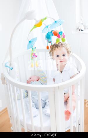 Süße lustige Kleinkind Mädchen sitzen in einer Krippe von ihr ihr neugeborenes Baby Bruder mit bunten Spielzeug und Baldachin Stockfoto