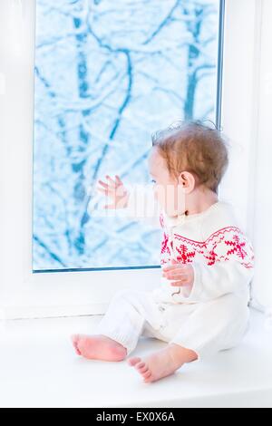 Glückliches lächelndes Baby sitzt am Fenster beobachten Schnee bedeckt Bäume im Garten Stockfoto