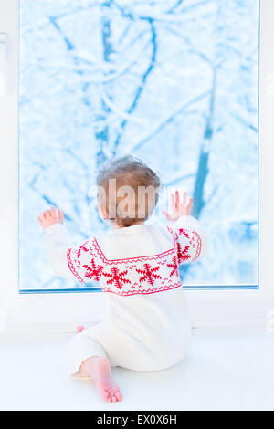 Glückliches lächelndes Baby sitzt am Fenster beobachten Schnee bedeckt Bäume im Garten Stockfoto