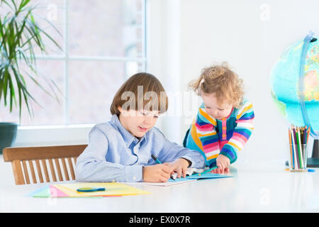 Junge Schuljunge macht seine Hausaufgaben und seine Kleinkind Schwester beobachtete ihn in einem weißen Raum neben einem Fenster Stockfoto