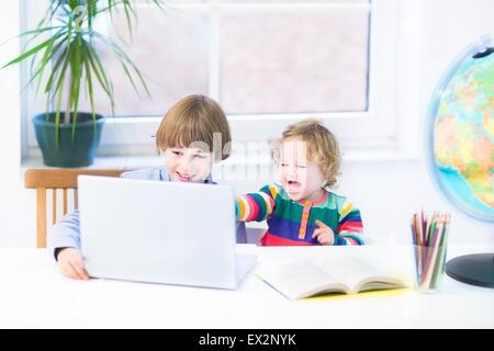 Zwei Kinder, Schuljunge und seine Schwester Kleinkind spielen zusammen mit einem Laptop an einen weißen Schreibtisch am Fenster sitzen Stockfoto