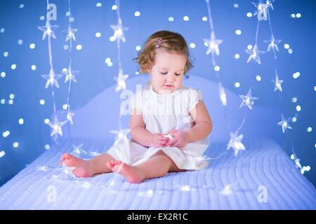 Schöne Kleinkind Mädchen mit dem lockigen Haar trägt ein weißes Kleid sitzt auf einem Bett zwischen Weihnachtsbeleuchtung Stockfoto