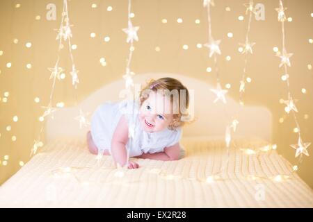 Schöne lockige Kleinkind Mädchen in einem weißen Kleid springen auf einem weißen Bett zwischen weichen warmen Weihnachtsbeleuchtung Stockfoto