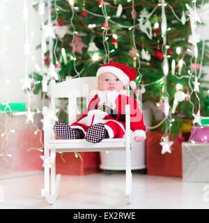 Lustige neugeborenes Baby gekleidet als Santa entspannen in einem weißen Schaukelstuhl unter einen schönen Weihnachtsbaum Stockfoto