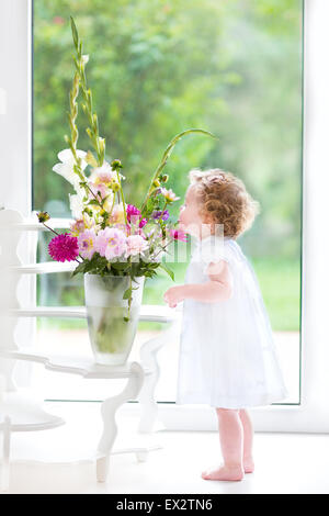 Niedlichen Kleinkind Mädchen riechen frische Blumen zu Hause neben ein großes Fenster mit Blick auf den Garten Stockfoto