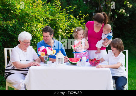 Große Familie mit Kindern haben Mittagessen im Freien. Eltern mit 3 Kindern und Großmutter Speisen im Garten. Stockfoto