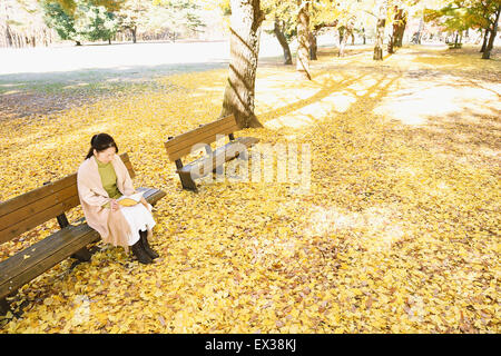 Ältere japanische Frau sitzen auf einer Bank mit einem Buch in einem Stadtpark im Herbst Stockfoto