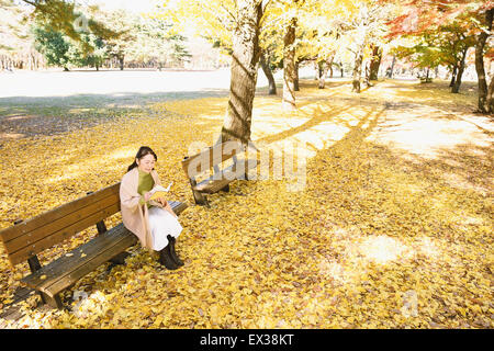 Ältere japanische Frau sitzen auf einer Bank mit einem Buch in einem Stadtpark im Herbst Stockfoto