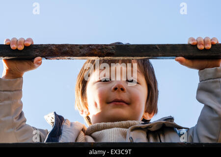 Kind, Junge, 4-5 Jahre alt, im Außenbereich. Das Tragen einer dicken Schicht und greifen eine Schiene, lächelnd und blickte nach unten im Viewer. Glücklichen Ausdruck. Low Angle View. Stockfoto