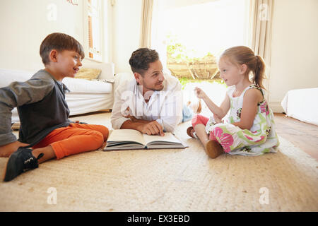 Familie mit Buch sitzen auf Boden mit kleinen Mädchen im Gespräch mit seinem Vater und Bruder. Vater mit seinem kleinen Jungen und Mädchen bei hom Stockfoto