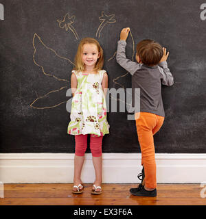 In voller Länge Portrait von niedliche kleine Mädchen stehen und kleiner Junge Engelsflügel um sie herum auf Tafel zeichnen. Stockfoto
