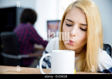 Junge Frau bläst auf einem Becher heißen Kaffee im Büro Stockfoto