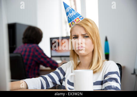 Traurige Frau mit Geburtstag Hut am Tisch im Büro sitzen und Blick in die Kamera Stockfoto