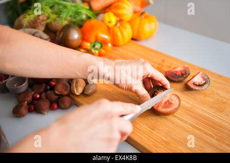 Zarte Frauenhand schneiden eine Tomate auf ein Holzbrett. Im Hintergrund, ein Haufen von Herbst Gemüse, einschließlich eine Paprika, Miniatur-Kürbisse, Kastanien, Preiselbeeren, Walnüsse und Pekannüsse. Stockfoto