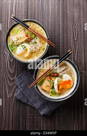 Asiatische Miso Ramen-Nudeln mit Ei, Tofu und Enoki in Schalen auf grauem Hintergrund aus Holz Stockfoto