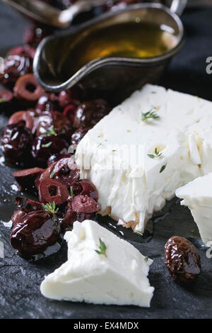 Ganze und geschnittene schwarze Oliven und Block von Feta Käse mit Olivenöl auf schwarzem Schiefer auf dunklem Hintergrund. Selektiven Fokus Stockfoto