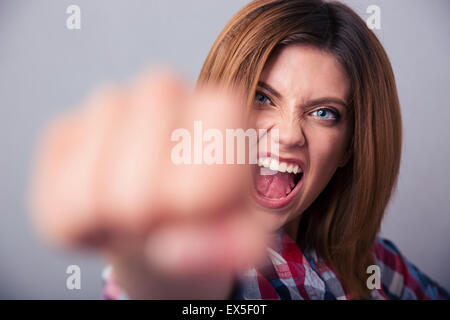 Böse Frau schlagen mit Faust vor der Kamera und schreien über grauen Hintergrund Stockfoto