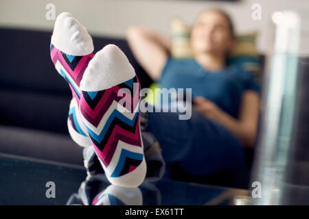 Kaukasische Mädchen Verlegung auf Sofa mit bunten Socken. Sie legt ihre Füße auf Tisch und entspannt. Das Mädchen sieht fern und hält Stockfoto