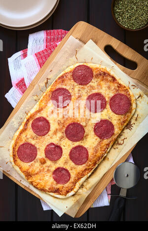 Hausgemachte Salami oder Salami Pizza auf Backpapier auf Holzbrett mit Tellern, getrockneter Oregano und Pizzaschneider auf der Seite Stockfoto