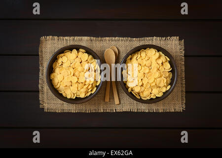 Knusprige Cornflakes Müsli in rustikale Schalen mit kleinen Holzlöffeln, fotografiert overhead auf dunklem Holz Stockfoto