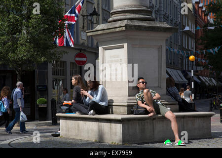 Sommer in London, England, Vereinigtes Königreich. Entspannen in der Sonne am Seven Dials in Covent Garden. Seven Dials ist eine kleine Kreuzung in Covent Garden im West End von London, wo sieben Straßen zusammenlaufen. Im Mittelpunkt der etwa kreisförmige Raum ist eine Säule Lager sechs Sonnenuhren, ein Ergebnis des Pfeilers vor eine späten Phase Änderung der Pläne von einer ursprünglichen sechs Straßen in Auftrag gegeben. Stockfoto