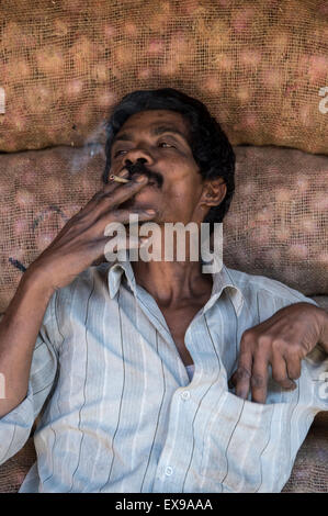 MYSORE, Indien - 4. November 2012: Indische Hersteller raucht eine Zigarette gegen Stacks Sackleinen Säcke auf dem Devaraja-Markt. Stockfoto