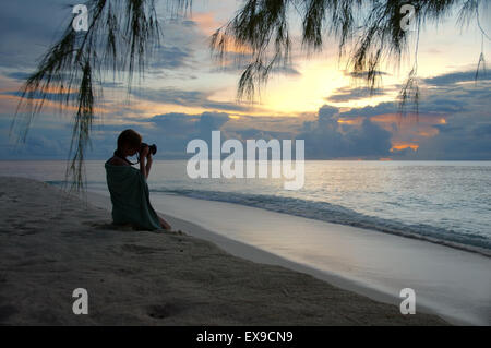 Eine junge Frau sitzt an den sandigen Ufern des Indischen Ozeans und fotografieren Sonnenuntergang, Denis Island, Seychellen Stockfoto