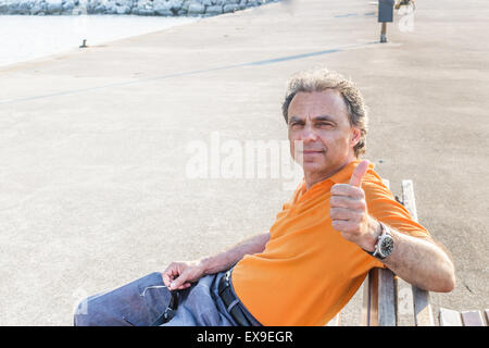 Edel senior Sportler mit drei-Tage-Bart und Salz und Pfeffer Haar eine orange Polo-Shirt trägt, während er auf einer Bank am Pier sitzt und Daumen auftauchen Stockfoto