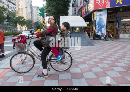 Asien, Taiwan, Taipei, Frau Reiten Fahrrad mit Sohn auf Rückseite entlang der geschäftigen Straßen der Stadt Stockfoto