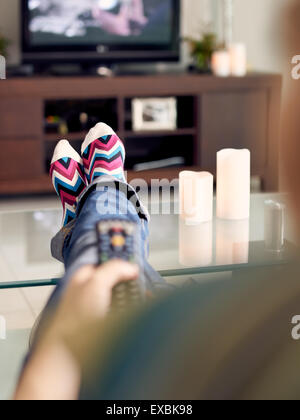 Kaukasische Mädchen Verlegung auf Sofa mit bunten Socken. Sie legt ihre Füße auf Tisch und entspannt. Das Mädchen sieht fern und hält Stockfoto