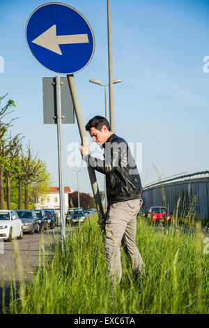 Junger Mann mit Lederjacke bewegen Pfeil Straßenschild in langen grünen Rasen Median neben Straße Stockfoto