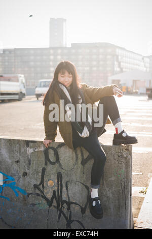 junge schöne asiatische Hipster Frau in das Stadtleben - Stadt während des Sonnenuntergangs in hinterleuchtet - sitzt auf einer Mauer mit smartphone Stockfoto