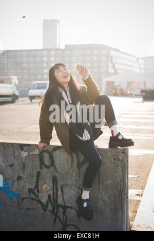 junge schöne asiatische Hipster Frau in das Stadtleben - Stadt während des Sonnenuntergangs in hinterleuchtet - sitzt auf einer Mauer mit smartphone Stockfoto