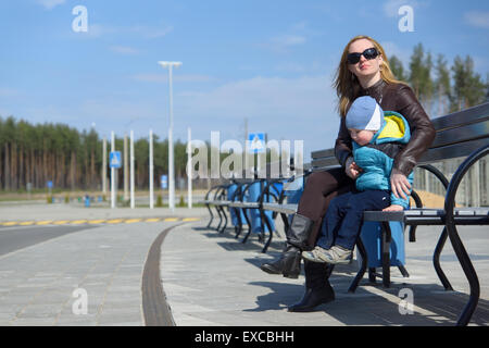 junge Mutter mit dem Sohn sitzt auf einer Bank unter dem blauen Himmel Stockfoto