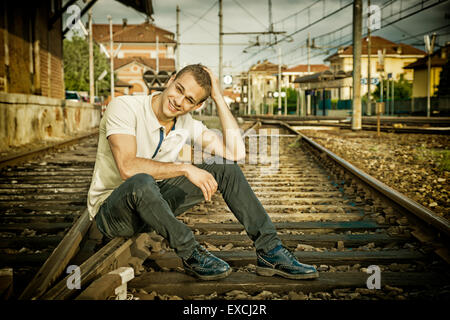 Attraktive junge Mann sitzt auf Eisenbahn, tragen gelbe Polo-Shirt und Jeans, Blick in die Kamera, Lächeln Stockfoto