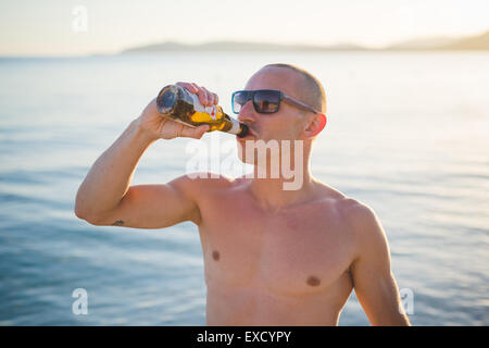 schönen jungen sportlichen Mann trinkt Bier am Strand Sonnenuntergang im Sommer Stockfoto