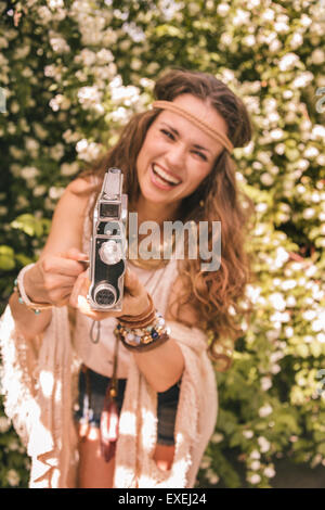 Langhaarigen Hippie aussehende junge Dame in gestrickten Schal und weiße Bluse ansehen unter den Blumen mit Retro-Kamera Stockfoto