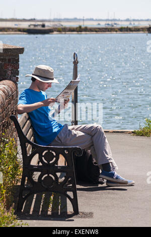 Junger Mann sitzt auf der Bank lesen Magazin am Hafen von Emsworth, Hampshire, UK im Juli Stockfoto