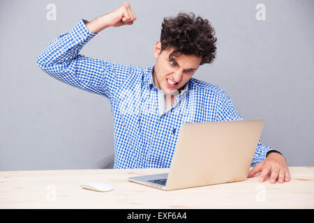 Verärgerter Mann sitzen am Tisch mit Laptop über grauen Hintergrund Stockfoto